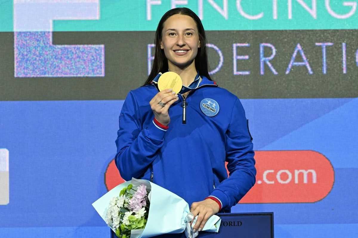 Athlete in blue jacket holding gold medal and bouquet on stage.