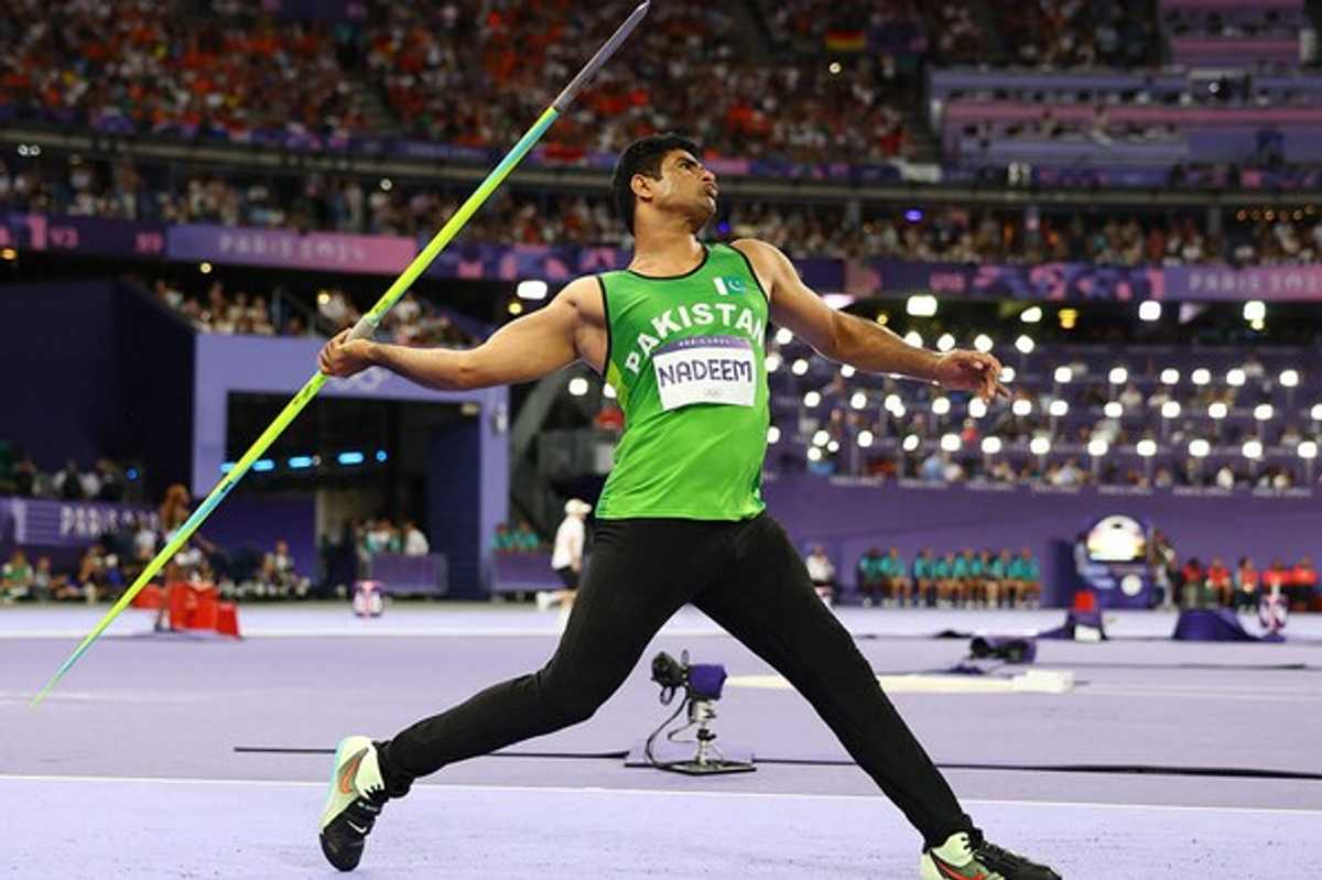 Athlete in green jersey throws javelin in stadium, audience in background.