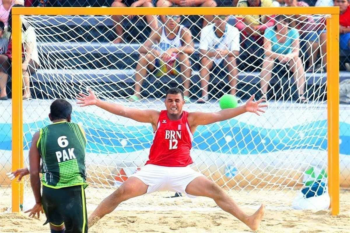 Beach handball goalie in red blocks a shot from opposing player in green, crowd watches.