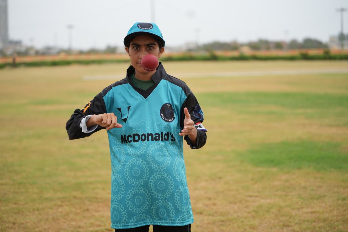 Cricket player balancing red ball on mouth on a grassy field.