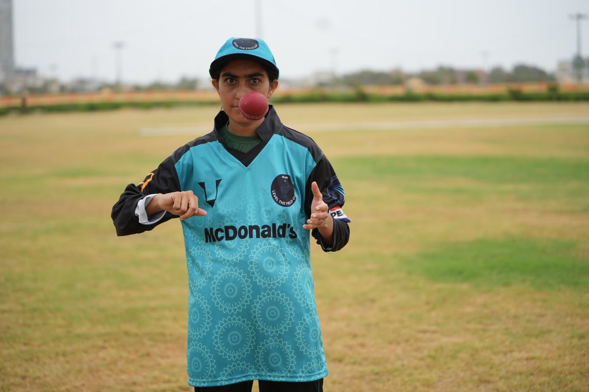Cricket player balancing red ball on mouth on a grassy field.