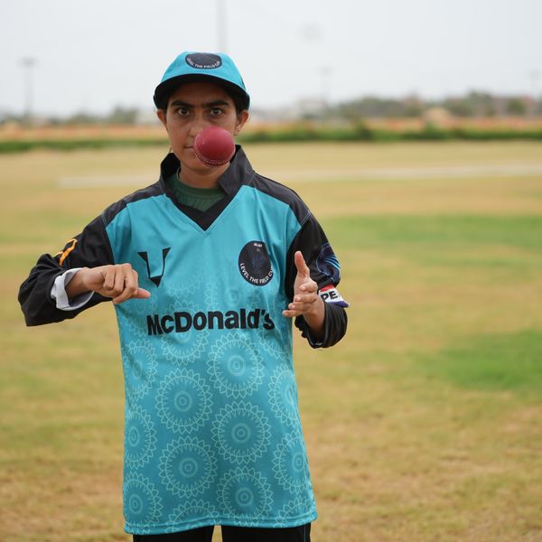 Cricket player balancing red ball on mouth on a grassy field.