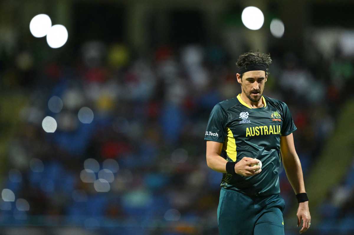 Cricketer in Australia kit holding a ball, blurred stadium crowd in background.