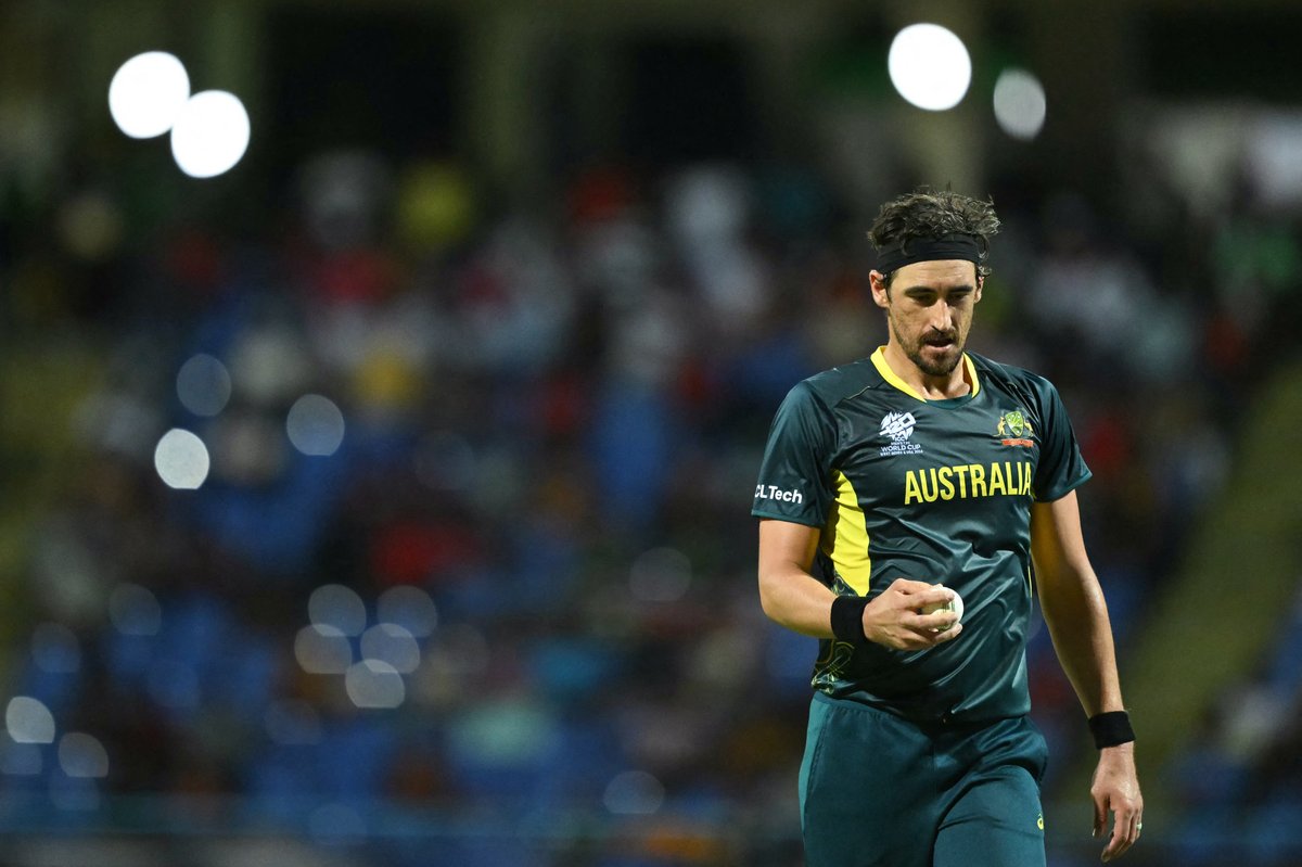 Cricketer in Australia kit holding a ball, blurred stadium crowd in background.