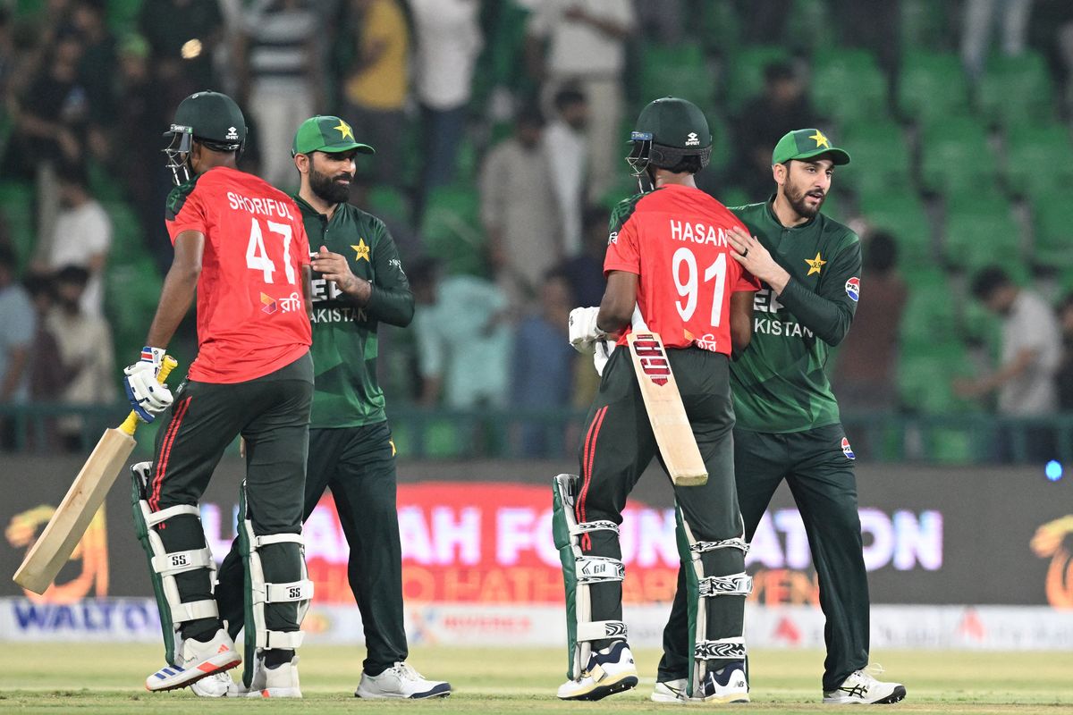 Cricketers shaking hands post-match, wearing green and red uniforms on a cricket field.