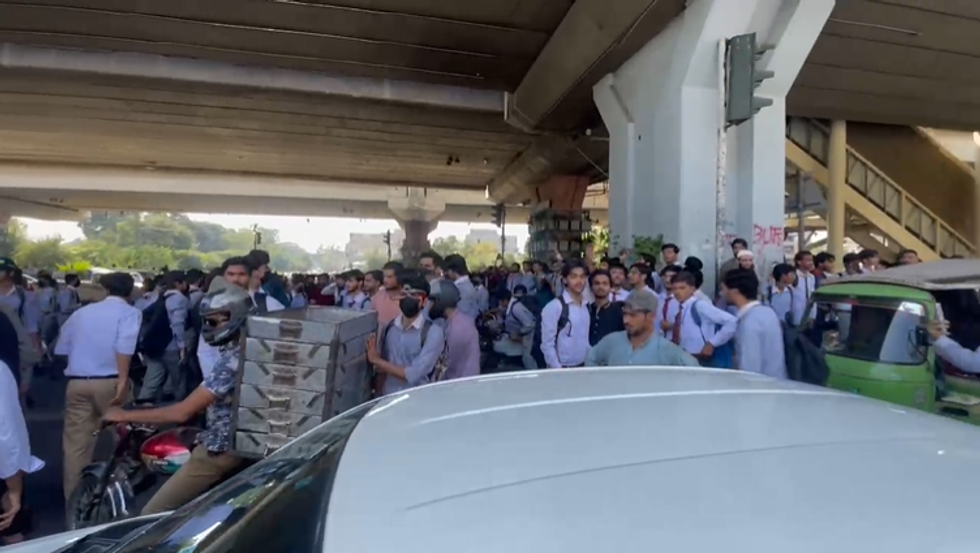 Crowd of people gathered under an overpass, with vehicles and trees in the background.