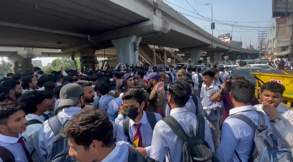 Crowd of students in uniforms gather under an overpass, with police and traffic nearby.