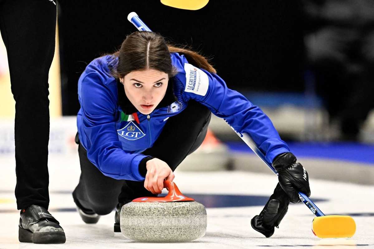 Curler in blue releasing a stone while teammate sweeps in a curling match.