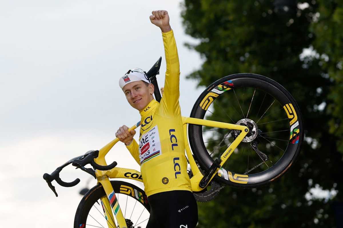 Cyclist in a yellow suit raises a bike triumphantly in front of trees.