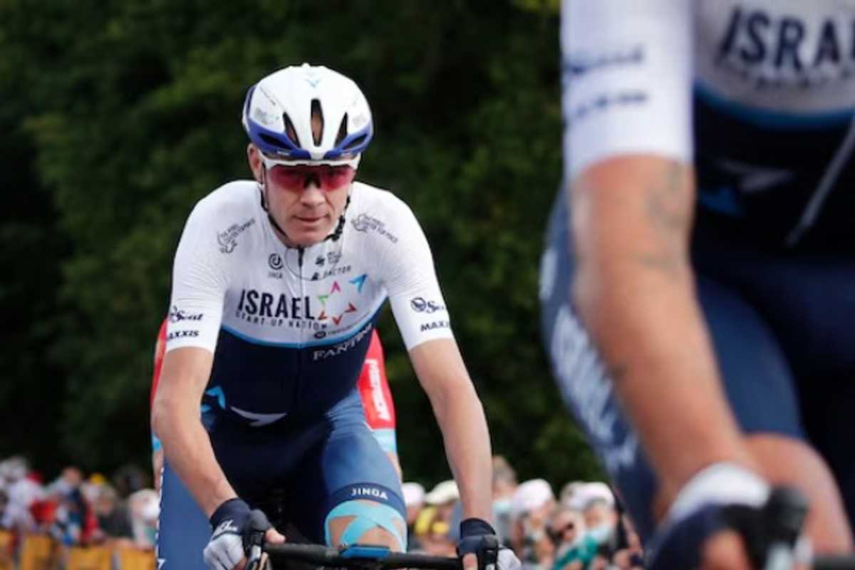 Cyclist in Israel team uniform riding in a race, with trees and spectators in the background.