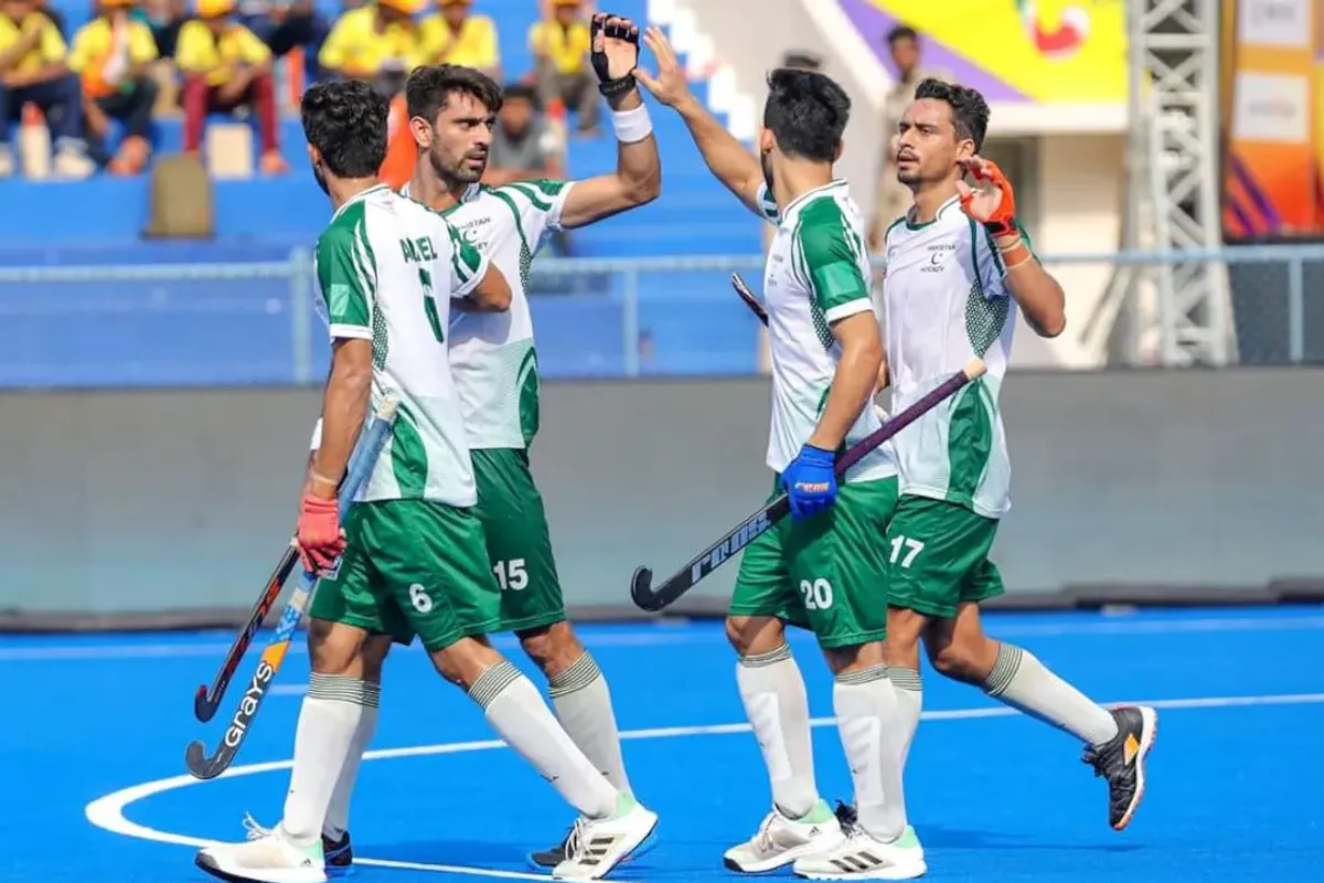 Field hockey players in green and white uniforms celebrate on a blue pitch.