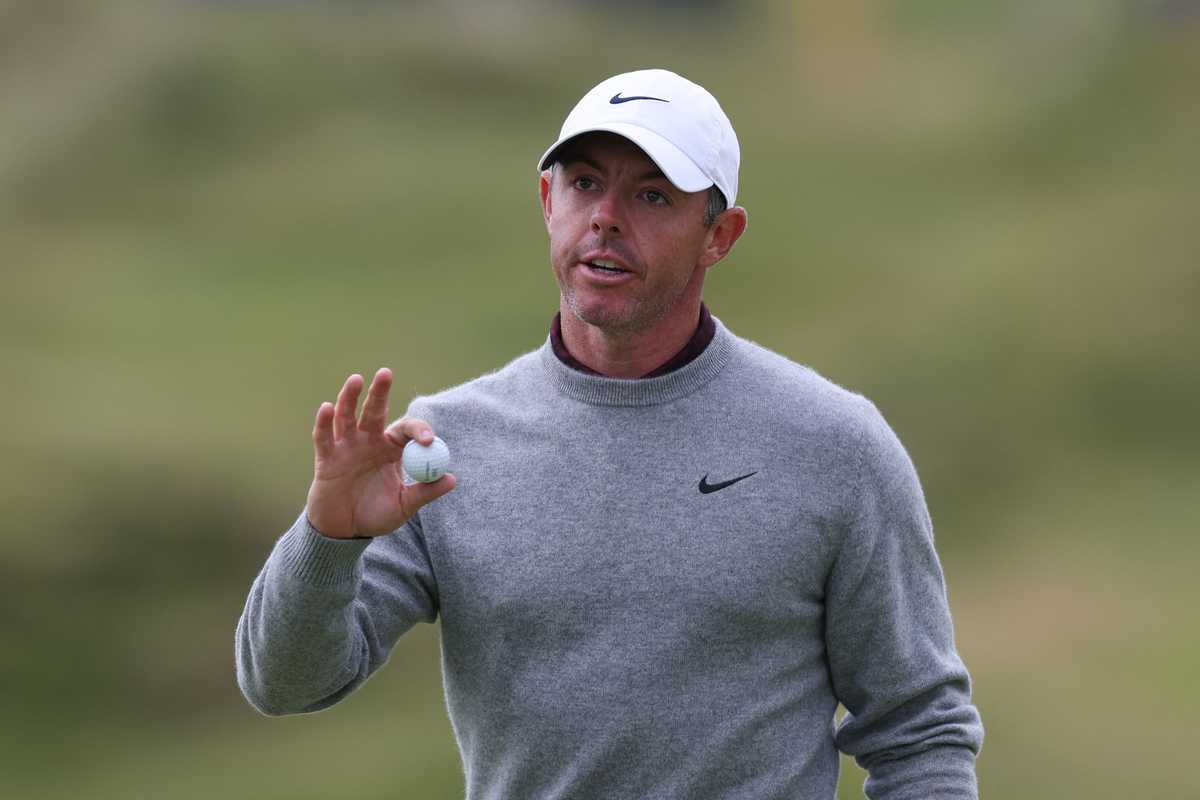 Golfer holding a ball, wearing a cap and gray sweater, on a golf course.