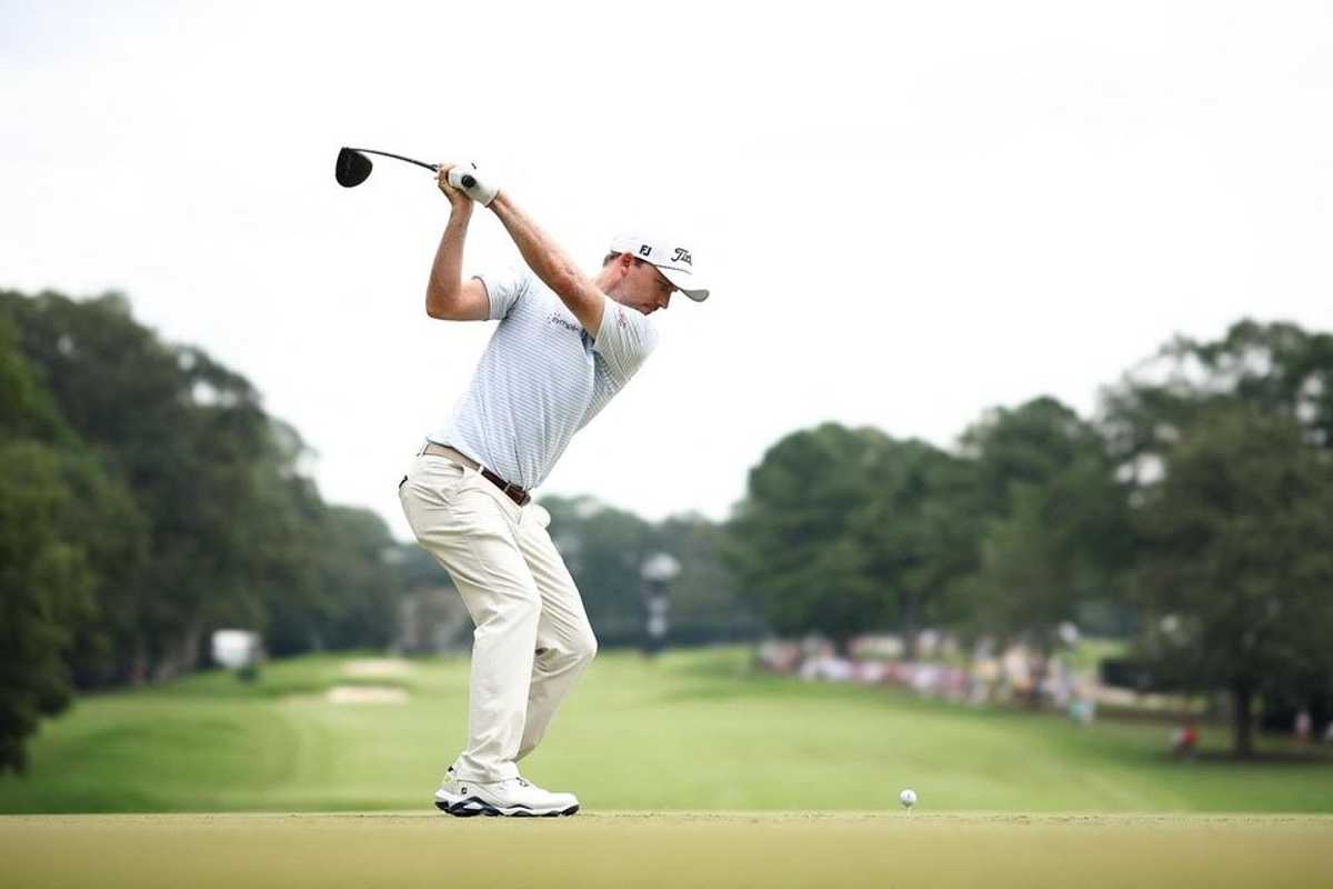 Golfer mid-swing on a lush course, ball on tee, trees in the background.