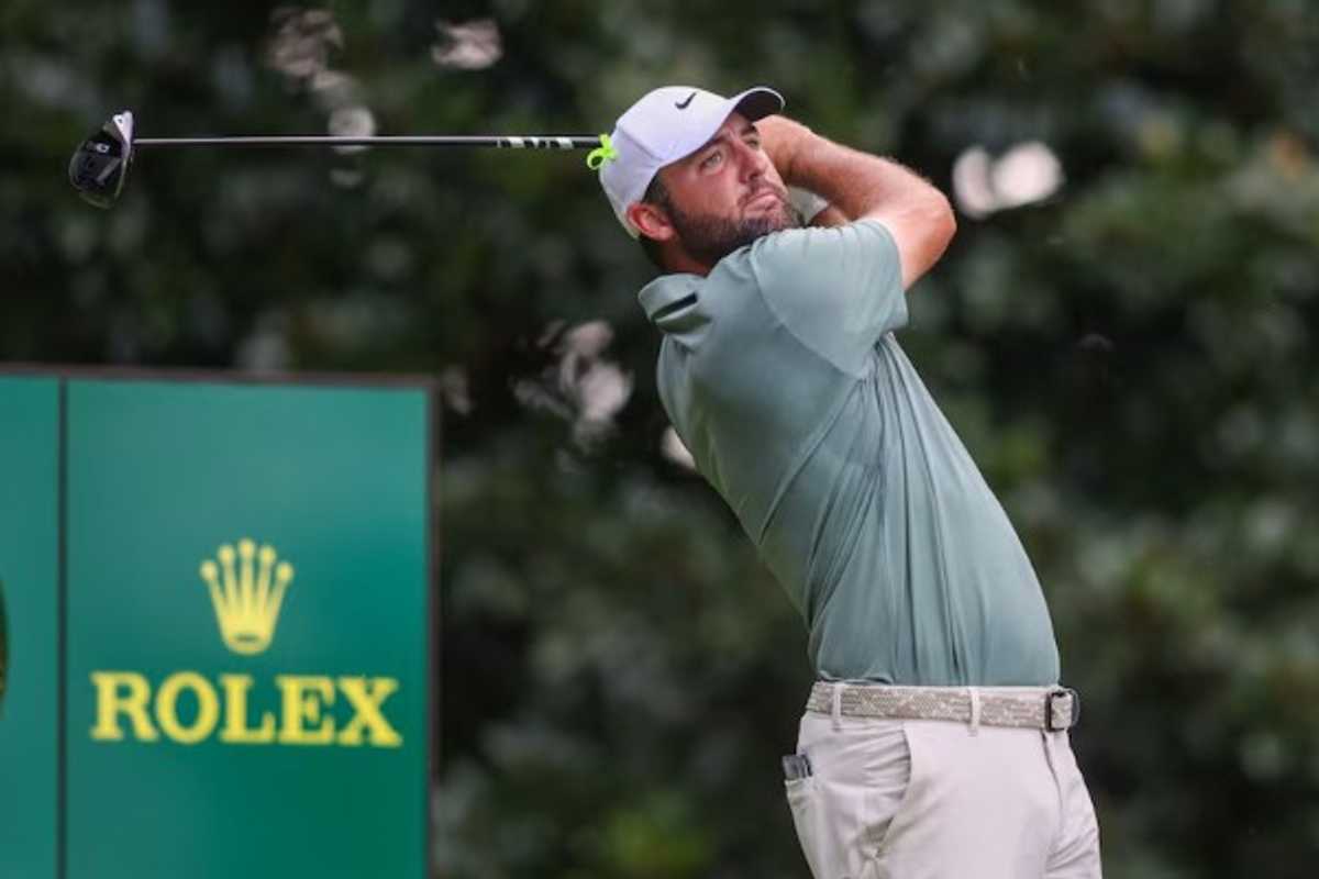 Golfer swings club near Rolex clock at a golf course, wearing a green shirt and white cap.