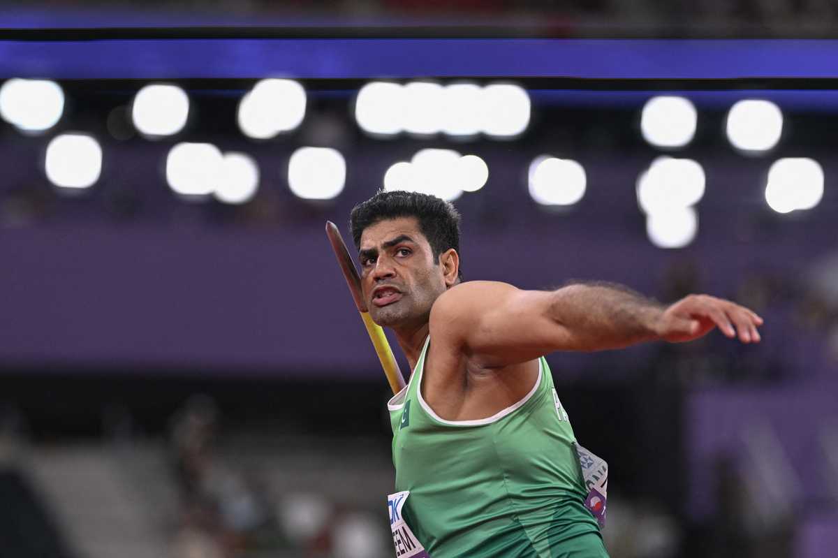 Javelin thrower in green uniform prepares to launch the javelin, stadium lights in background.