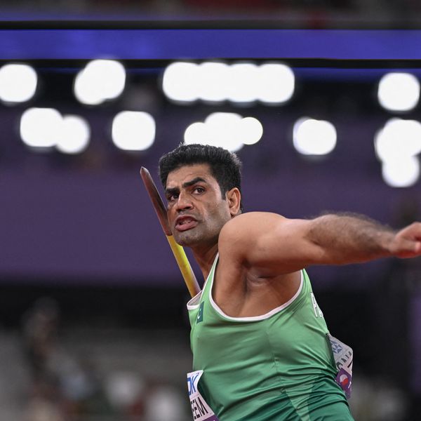 Javelin thrower in green uniform prepares to launch the javelin, stadium lights in background.