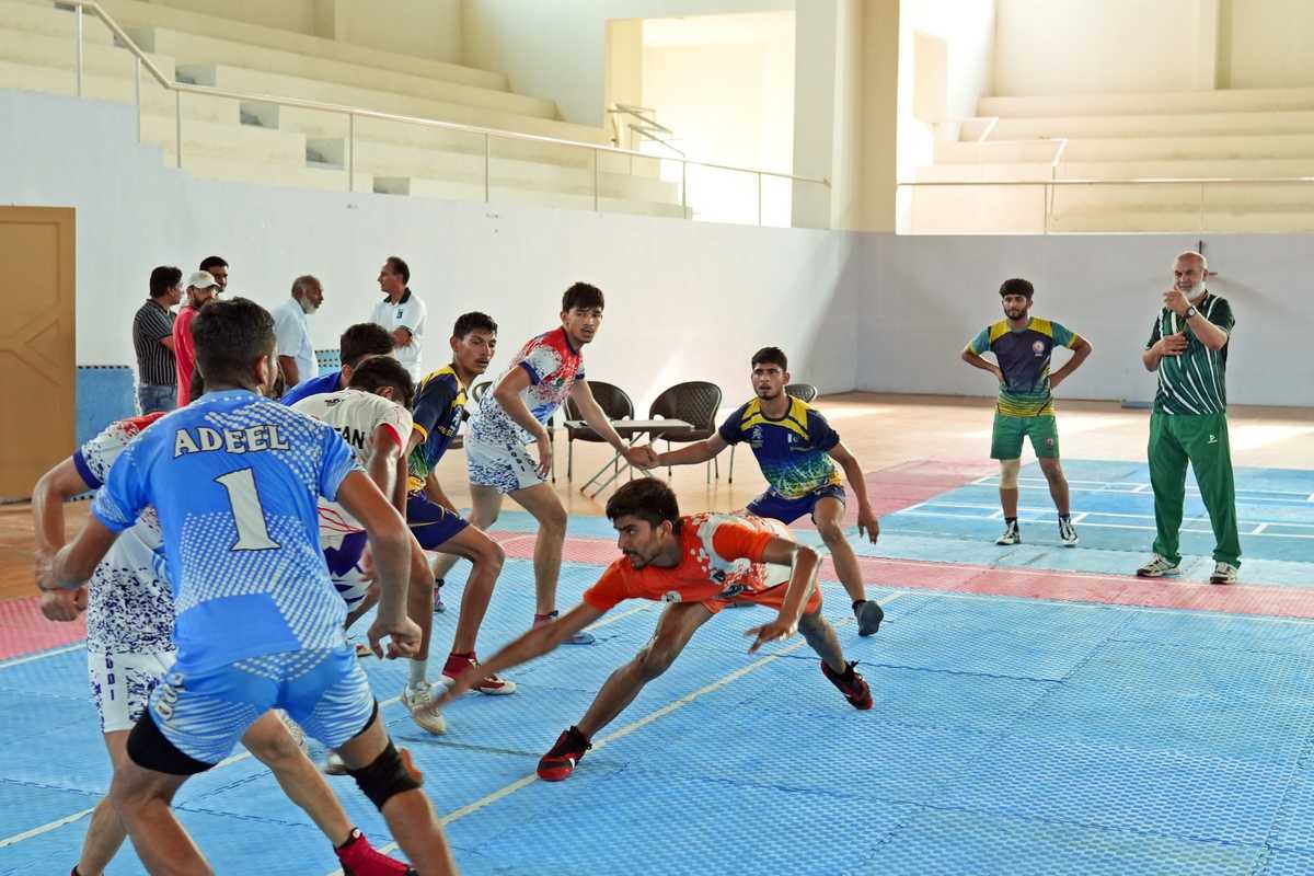 Kabaddi players in action on a blue mat, with a referee observing in the background.