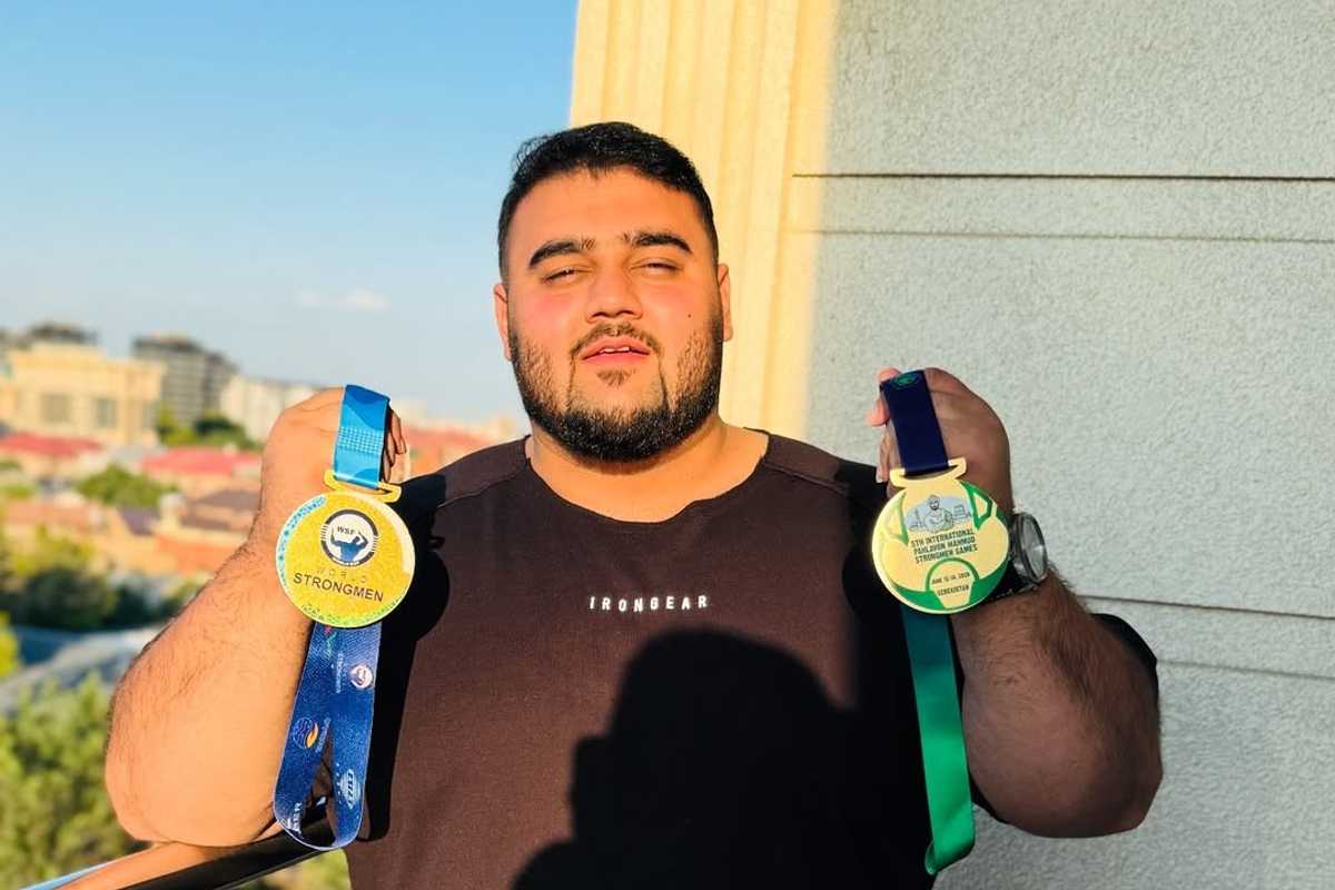 Man holding two Strongmen competition medals, standing outdoors in sunlight.