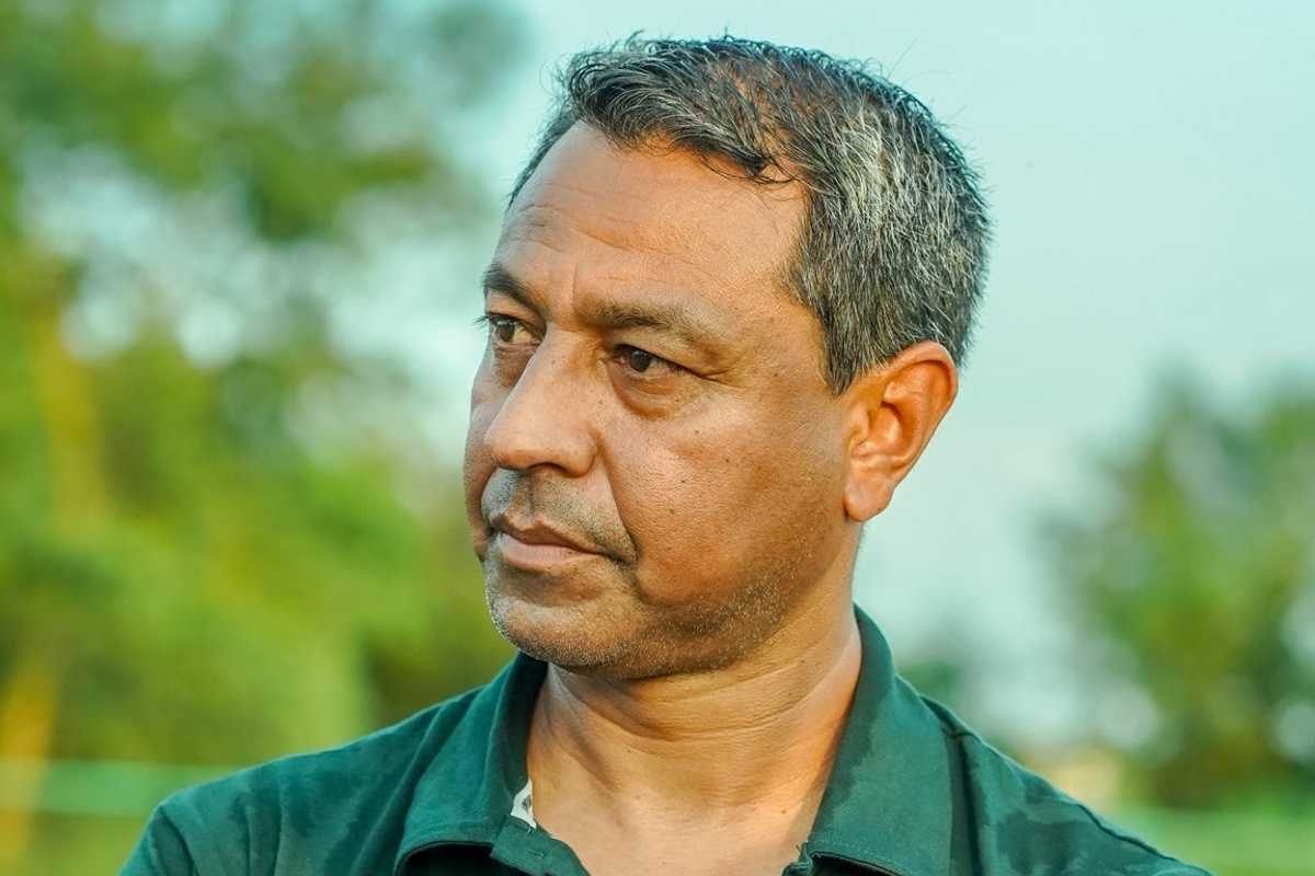 Man in a green shirt pensively gazing to the side, with blurred greenery in the background.