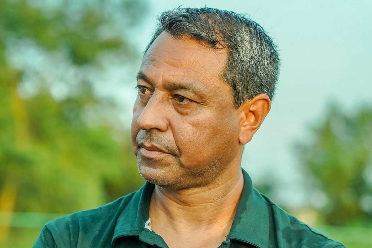 Man in a green shirt pensively gazing to the side, with blurred greenery in the background.