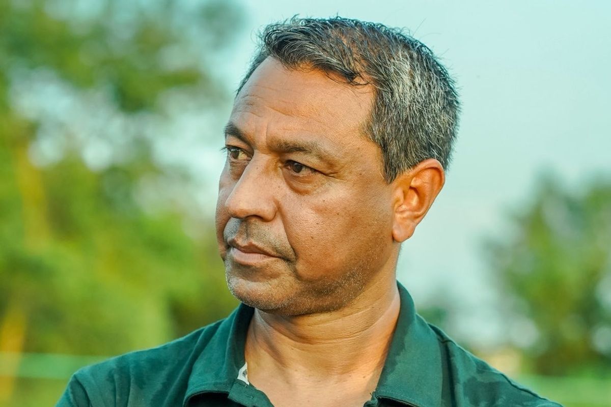 Man in a green shirt pensively gazing to the side, with blurred greenery in the background.