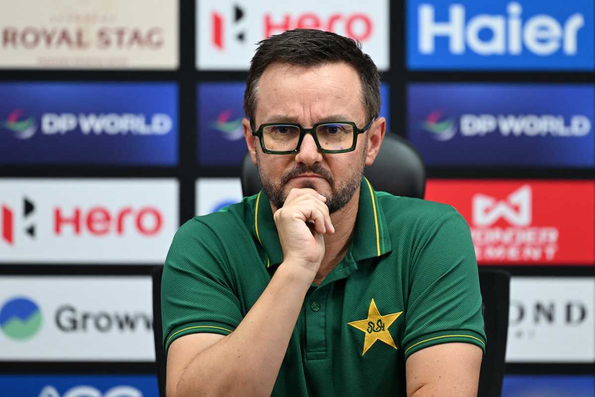Man in a green shirt with star logo, sitting pensively at a press conference.