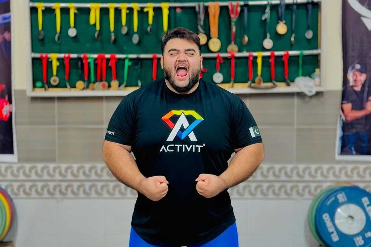 Man in a gym flexing muscles, medals displayed behind him.