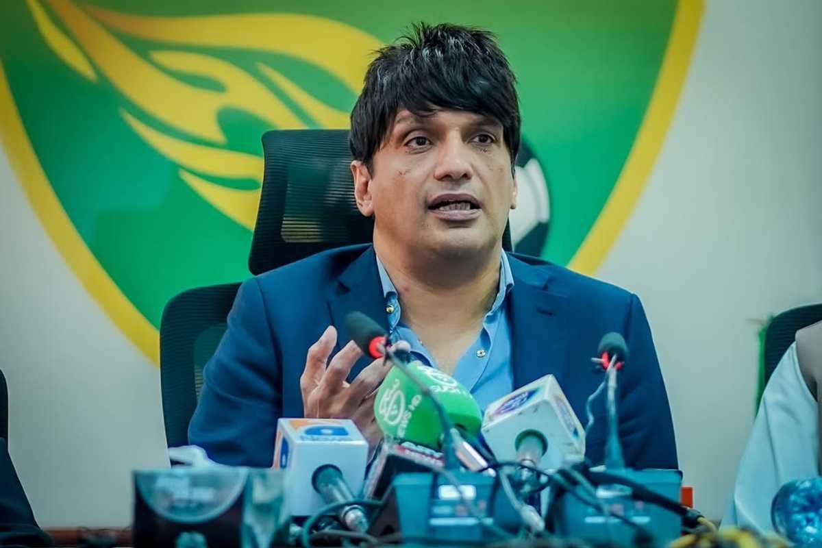 Man in a suit speaking at a press conference, surrounded by microphones.