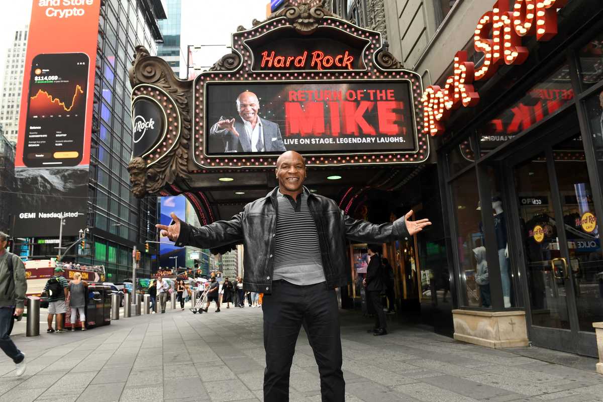 Man standing with arms outstretched in front of Hard Rock Cafe marquee in a city.