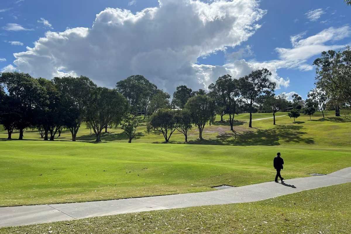 Man walking on path through a sunny park with trees and large clouds above.