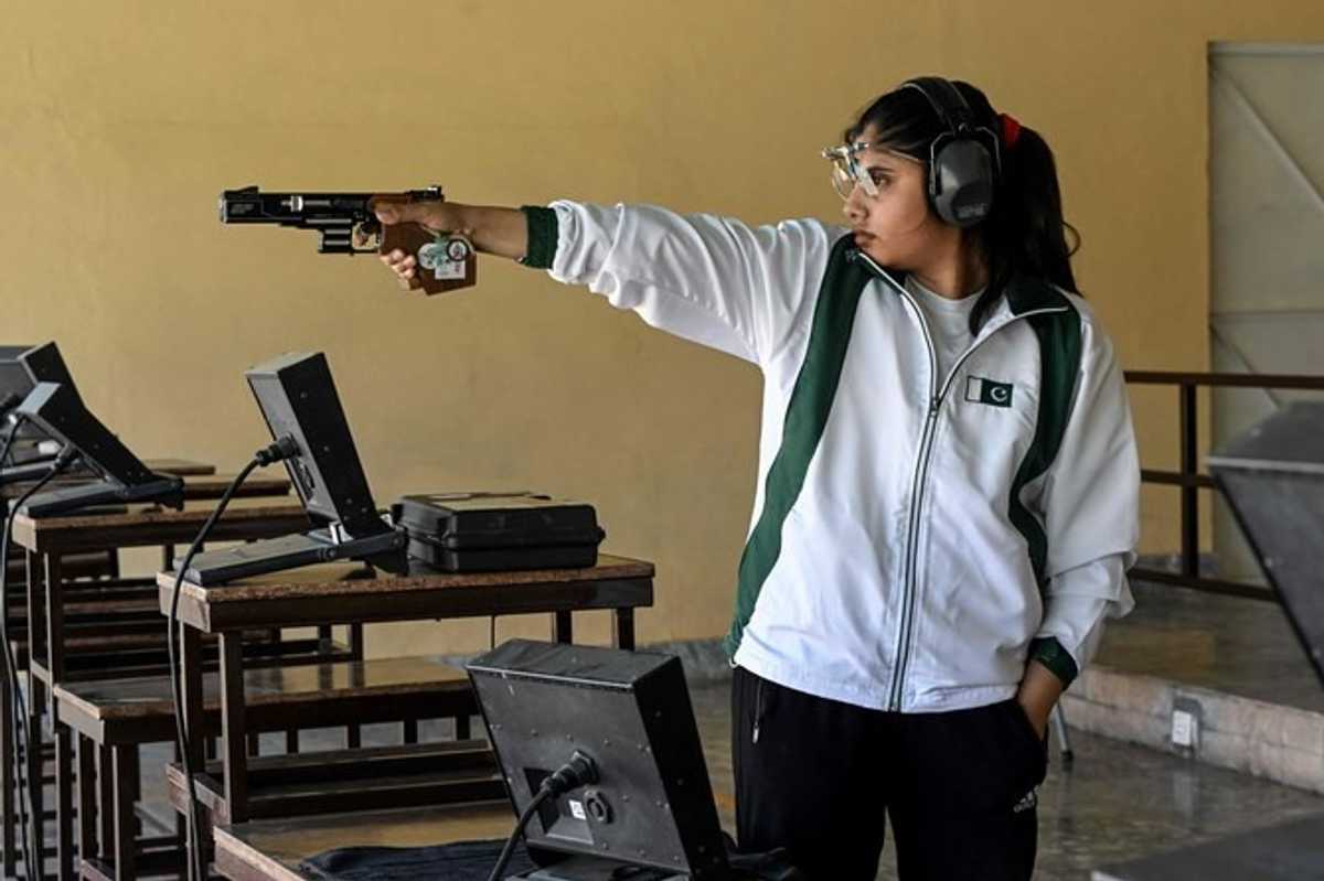 Person in a tracksuit aiming a pistol at an indoor shooting range, wearing ear protection.