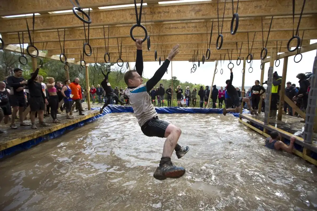 Person swings on rings over a muddy obstacle course with spectators watching.