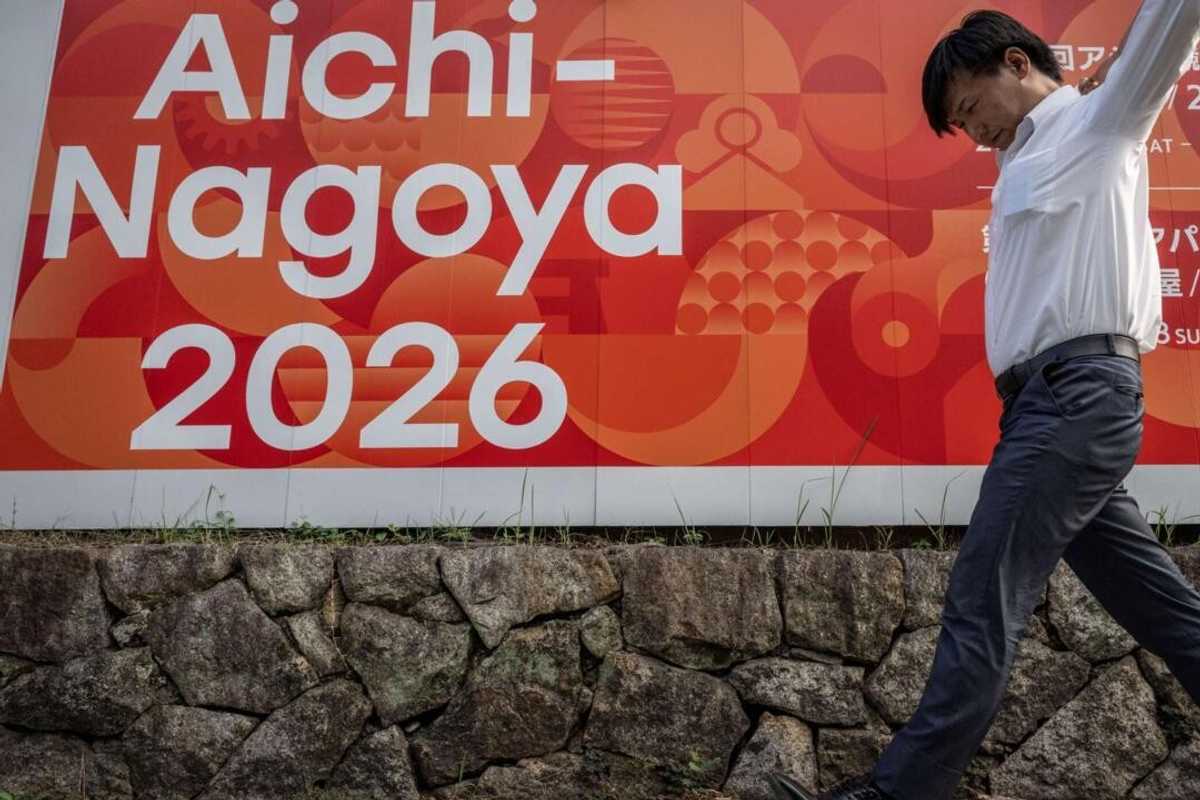 Person walking past an "Aichi-Nagoya 2026" sign with an abstract red background.