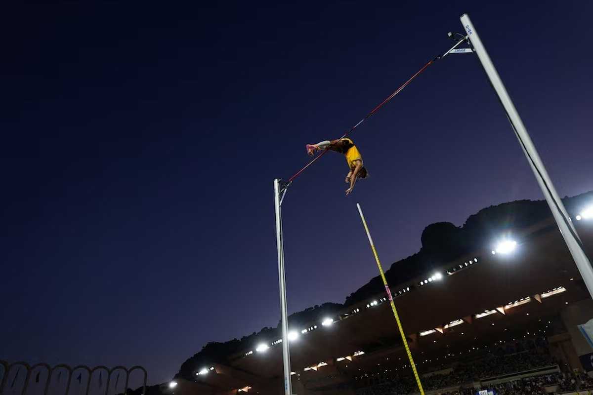 Pole vaulter clearing bar under a twilight sky in a stadium.