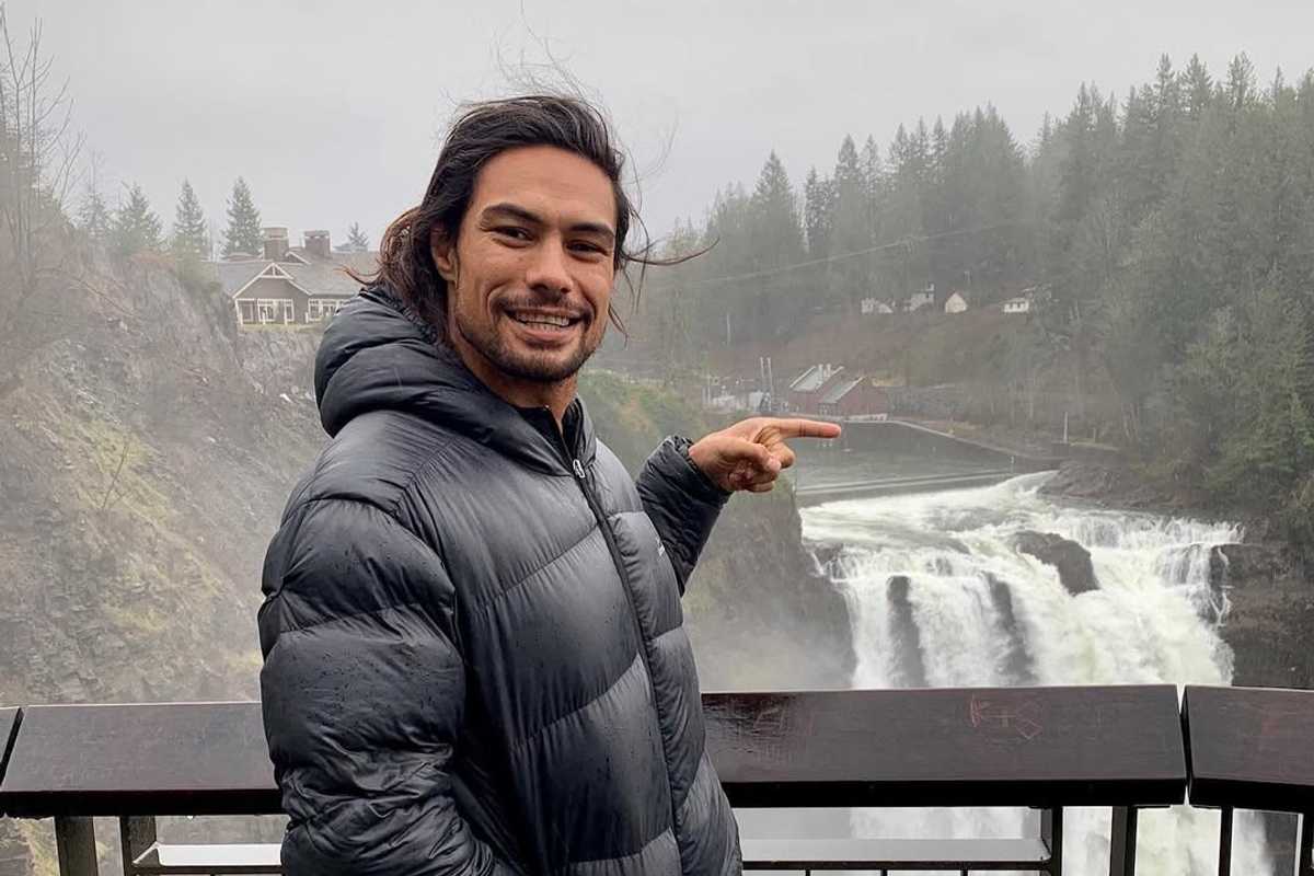 Smiling man in a puffer jacket points at a waterfall in a misty forest setting.
