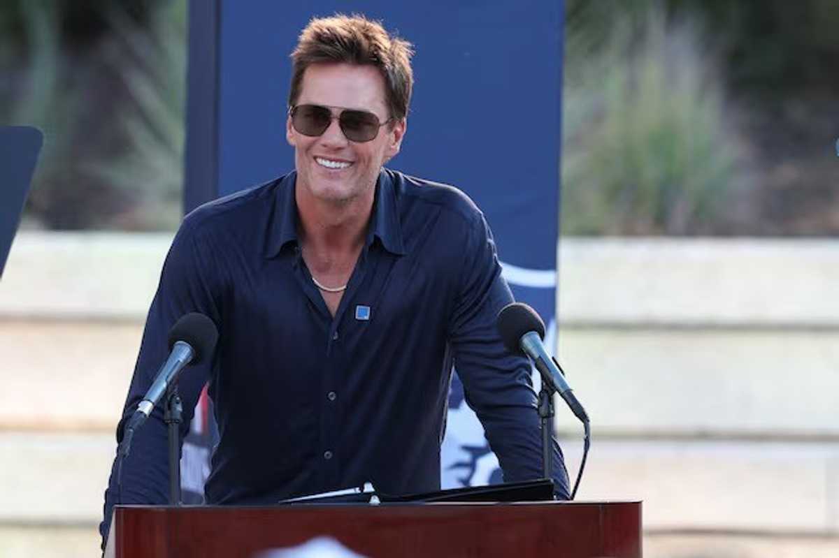 Smiling man in sunglasses speaking at a podium with two microphones outdoors.