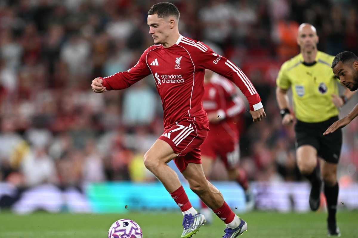 Soccer player in red kit dribbles ball during a match, with a referee and player nearby.