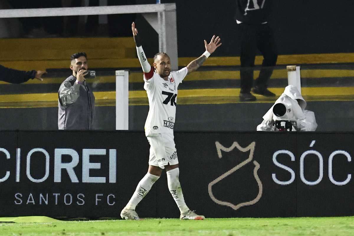 Soccer player in white kit celebrates with arms raised, fans and camera in background.