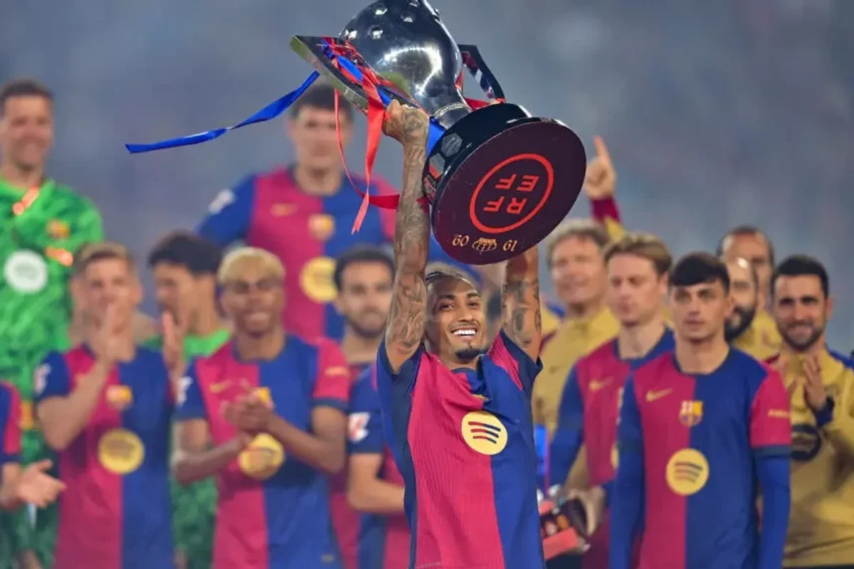 Soccer player lifts a trophy, teammates in Barcelona kits applaud in the background.