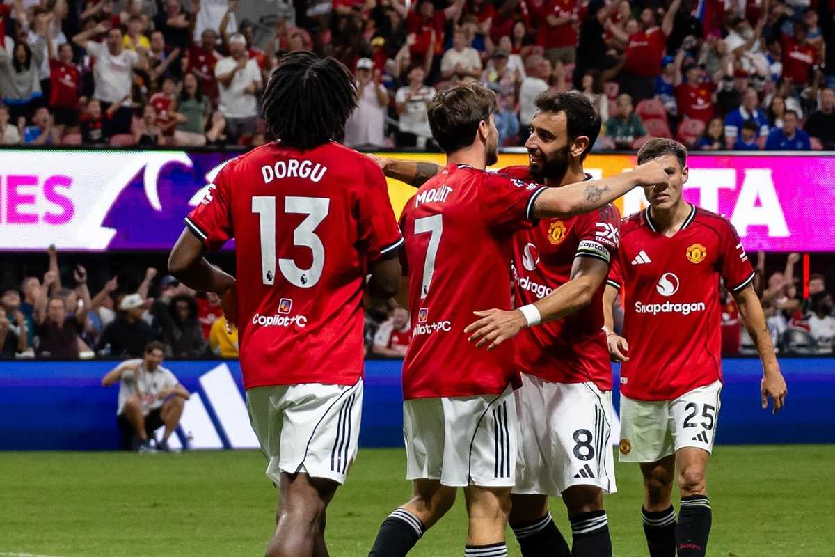 Soccer players in red jerseys celebrate on the field with cheering crowd behind them.