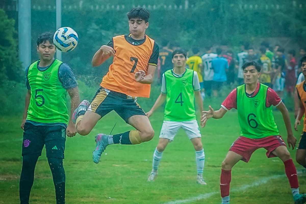Soccer players in training, one mid-air, on a grassy field wearing colorful vests.