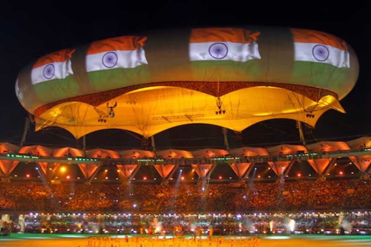 Stadium at night with glowing canopy and Indian flags projected above a crowd.