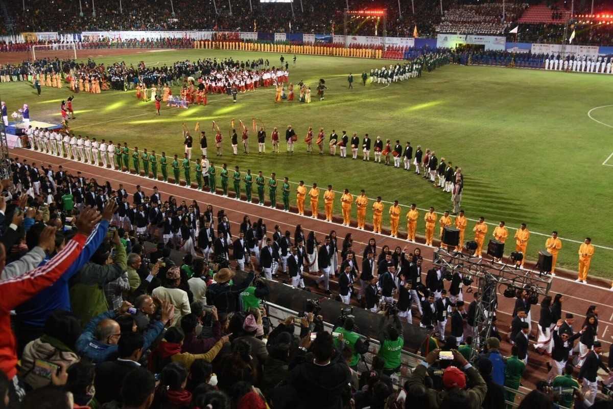 Stadium ceremony with performers in colorful uniforms, crowded stands, and vibrant lighting.