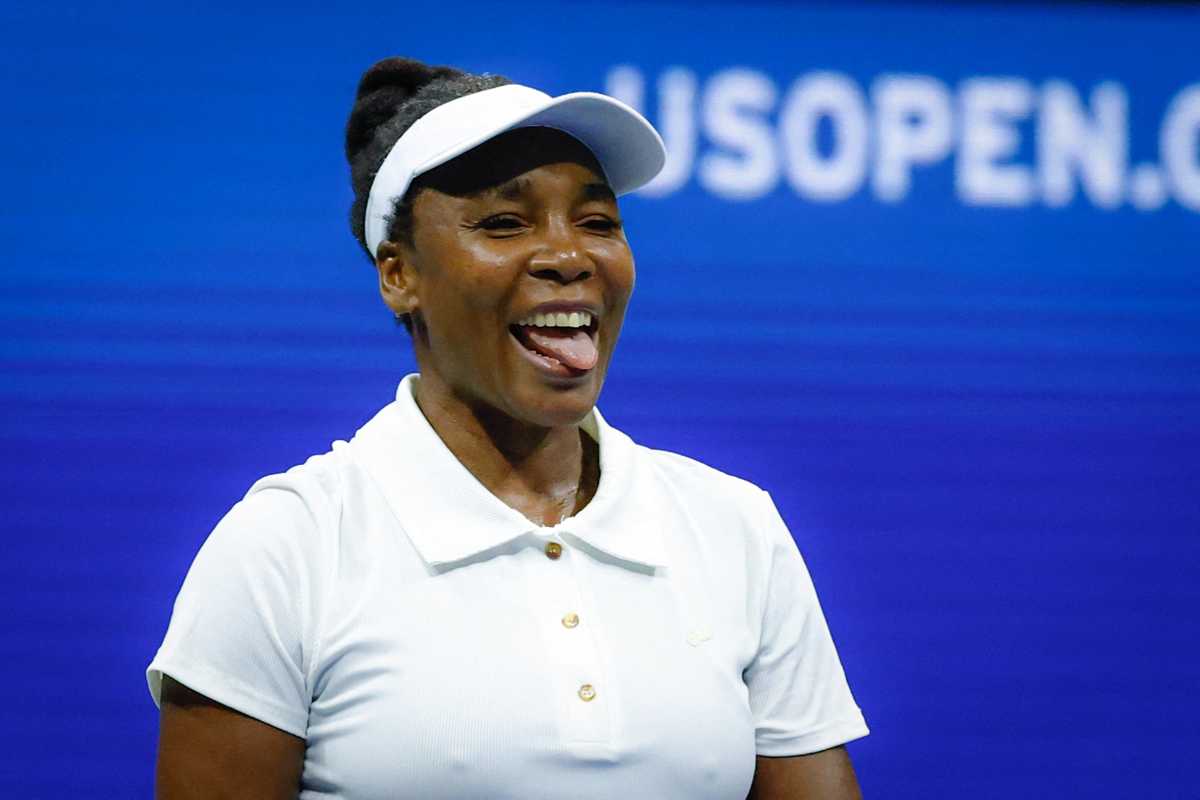 Tennis player smiling with tongue out, wearing a white shirt and visor, US Open background.