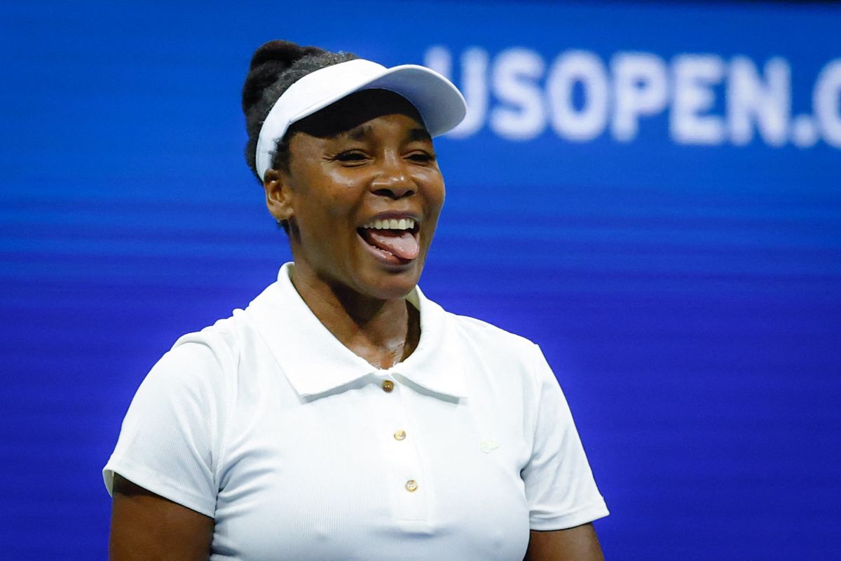 Tennis player smiling with tongue out, wearing a white shirt and visor, US Open background.