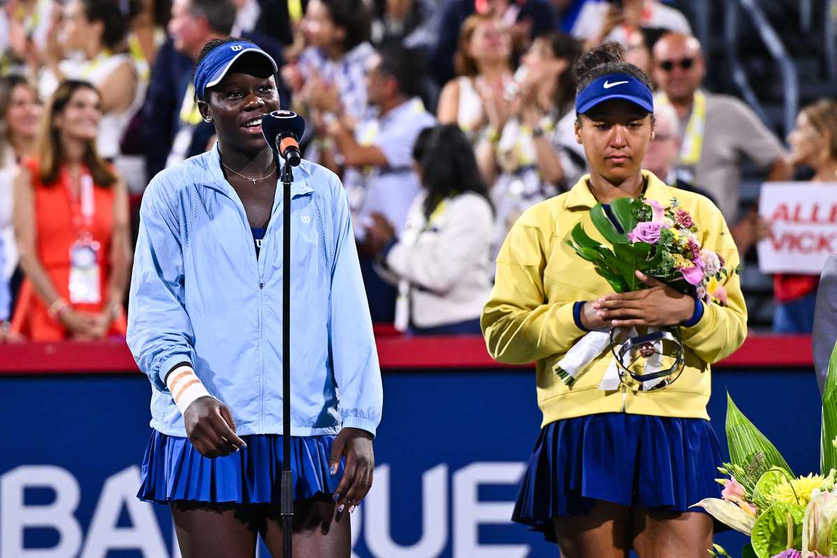 Tennis players at a podium; one speaks, the other holds flowers, surrounded by applauding crowd.