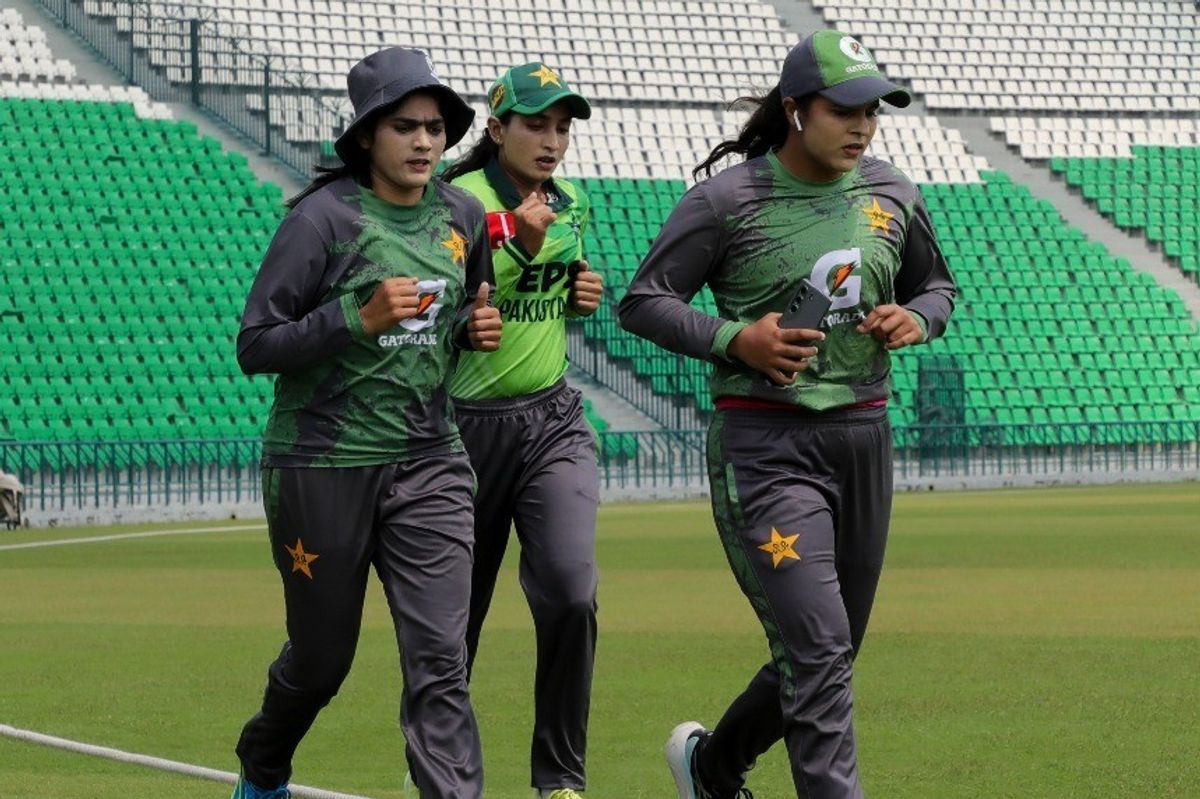 Three female cricketers jogging on a field in green and gray uniforms.