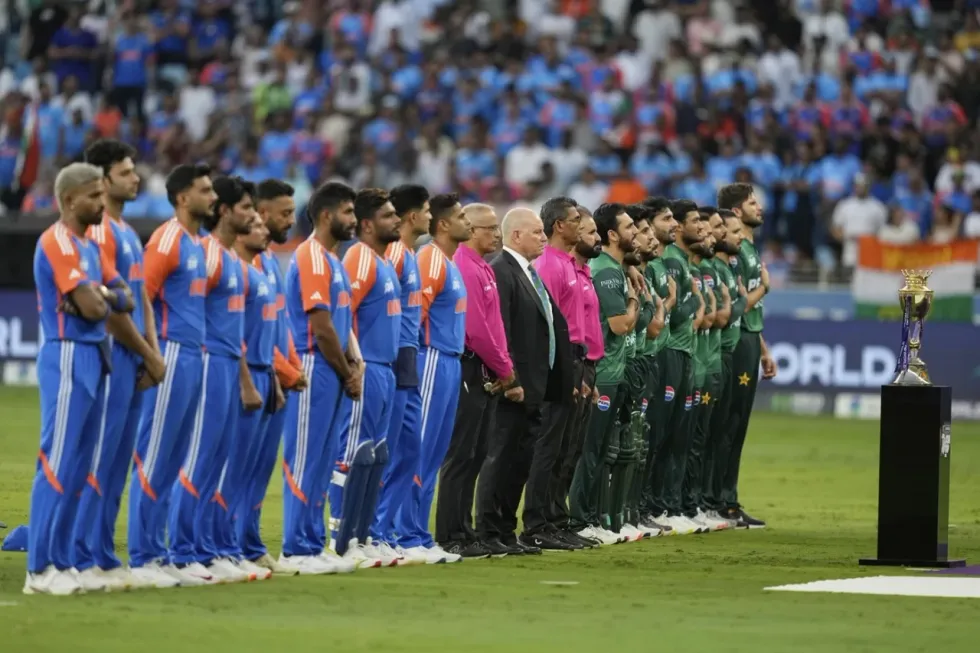 Two cricket teams stand facing a trophy on a field, with a crowd in the background.
