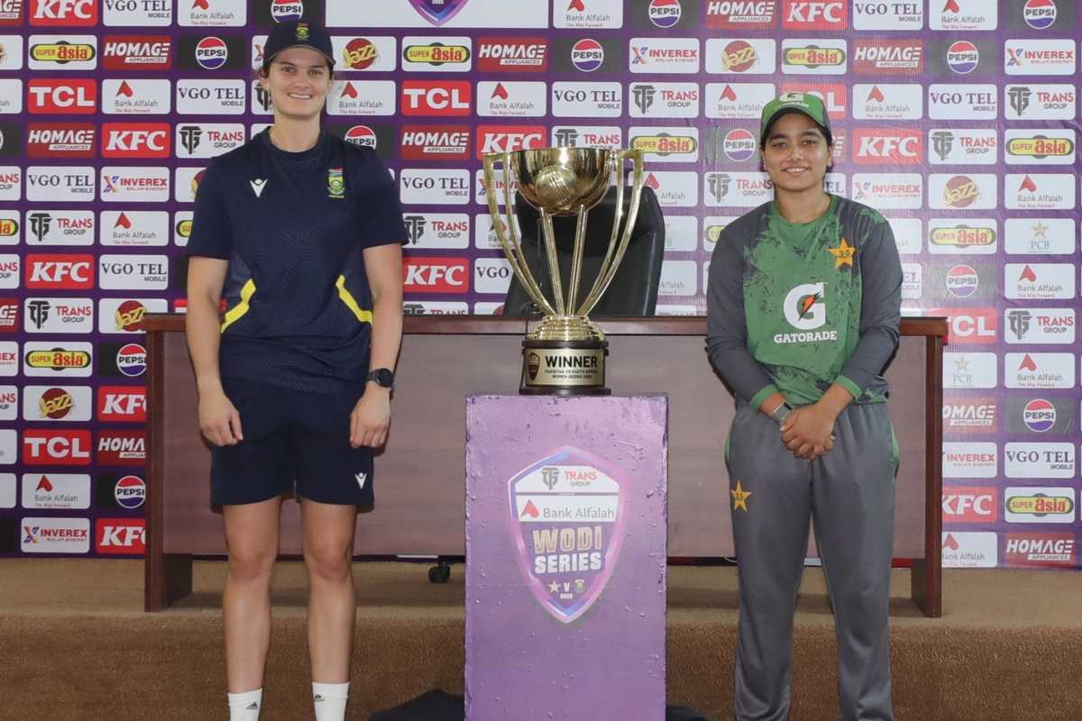 Two cricketers stand beside a trophy on a podium at a press event.