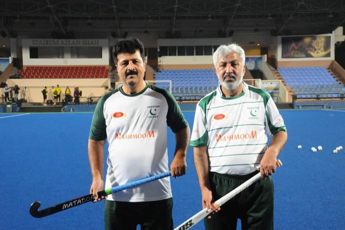 Two men holding hockey sticks on a blue field, wearing Pakistan team uniforms.